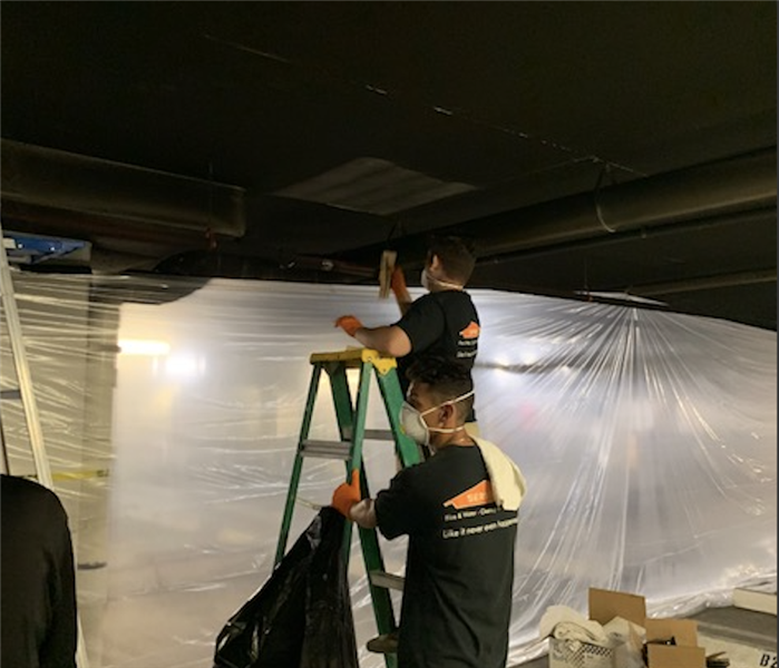 Two SERVPRO employees wearing protective gloves and mask, one on a ladder cleaning smoke damage and other man holding ladder